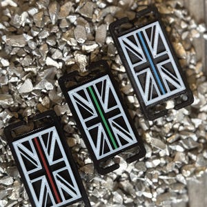 May include: Three black keychains with a Union Jack design. Each keychain has a different coloured stripe: red, green, and blue. The keychains are displayed on a bed of small, grey stones.