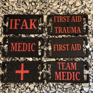 May include: Six black tactical patches with red text, including "IFAK", "FIRST AID TRAUMA", "MEDIC", "FIRST AID", a red cross, and "TEAM MEDIC". The patches are displayed on a background of small, grey stones.
