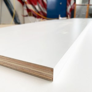 Puede incluir: Un panel de madera rectangular blanco con una superficie lisa y brillante. El borde revela la construcci&oacute;n en capas de la madera, mostrando la veta y la textura naturales. El panel es probablemente para construcci&oacute;n o fabricaci&oacute;n de muebles.