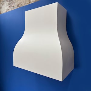 May include: A white range hood with a curved design, mounted on a blue wall. The hood has a rectangular top and a wider, flared bottom. The background includes a section of exposed brick.