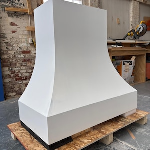 May include: A large, white range hood with a curved design, resting on a wooden pallet. The hood has a rectangular base and a flared top, showcasing a modern aesthetic. The background includes a workshop setting with tools and a window.