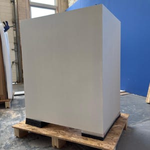 May include: A large, rectangular, off-white structure, possibly a display or storage unit, sits atop a wooden pallet. The unit is elevated by black foam pads. The background includes a blue wall and a window.