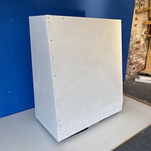 May include: A white rectangular cabinet with a slightly angled top. The cabinet is constructed with visible screws along the edges. It sits on a light gray surface against a blue wall.