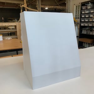 May include: A white, angular, modern-style range hood. The hood has a slanted front and a rectangular base. The structure is made of a solid, opaque material. The background shows a workshop setting with tools and equipment.