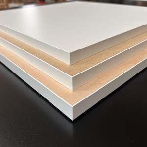 Puede incluir: Primer plano de tres paneles rectangulares apilados. Cada panel tiene una superficie superior blanca y un n&uacute;cleo de color marr&oacute;n claro, similar a la madera. Los bordes est&aacute;n acabados con un borde blanco. Los paneles est&aacute;n sobre una superficie oscura.