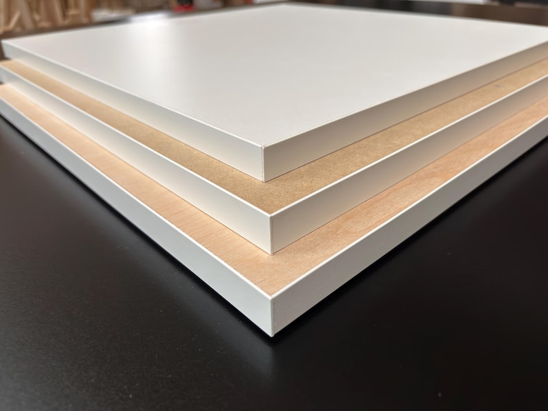 Puede incluir: Primer plano de tres paneles rectangulares apilados. Cada panel tiene una superficie superior blanca y un n&uacute;cleo de color marr&oacute;n claro, similar a la madera. Los bordes est&aacute;n acabados con un borde blanco. Los paneles est&aacute;n sobre una superficie oscura.