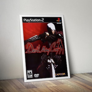 May include: Framed poster for the PlayStation 2 game, "Devil May Cry." The artwork features a character in a dynamic pose against a red background. The game's title is prominently displayed in a stylized font. The poster is in a white frame.