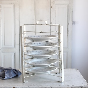 May include: A white metal plate rack holding six white plates with blue floral designs. The rack is on a white table, with a blue cloth on the left. The background features white doors.