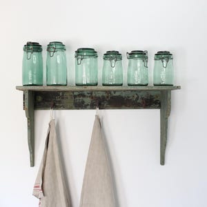 Op de afbeelding: Zes vintage aqua glazen potten met metalen sluitingen staan op een verweerde groene plank. Twee beige linnen handdoeken hangen eronder. De scène roept een rustieke, boerderijstijl op.