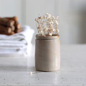 Puede incluir: Un pequeño jarrón de cerámica beige lleno de flores blancas secas. El jarrón está sobre una superficie de madera blanca desgastada. En el fondo, un manojo de palitos de canela atados con hilo descansa sobre una pila de toallas blancas.