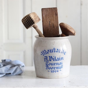 Op de afbeelding: Een vintage keramische mosterdpot met blauwe letters en houten keukengereedschap. Op de pot staat "Moutarde A. Vilain Tournai 1864". De houten gereedschappen omvatten een vleeshamer, een boterspaan en een klein houten deksel.