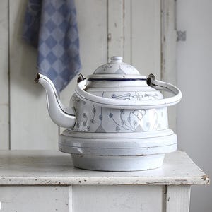 May include: A white enamel teapot featuring a delicate grey floral pattern and a matching lid. The teapot has a curved spout, a rounded handle, and a wide base. The metal surface shows signs of wear, including small chips on the spout and handle.