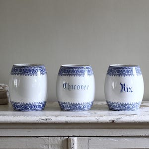 Op de afbeelding: Drie witte keramische voorraadpotten in de vorm van een ton, voorzien van blauwe bloemmotieven aan de boven- en onderrand. Twee van de potten hebben blauwe tekst op de voorzijde, Chicoree en Riz, terwijl de derde pot blanco is.