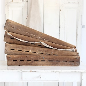 May include: A stack of four rectangular wooden crates with weathered brown surfaces. Each crate features a series of horizontal ventilation slots along the side panels. A piece of off-white fabric is draped between the top two wooden crates.