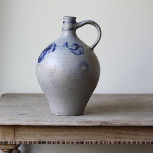 Pode incluir: Um jarro de grés cinza-claro com corpo bulboso e uma única alça. O recipiente apresenta motivos florais azul-cobalto pintados à mão ao redor do ombro e faixas azuis horizontais ao redor do pescoço estreito, com um acabamento fosco.