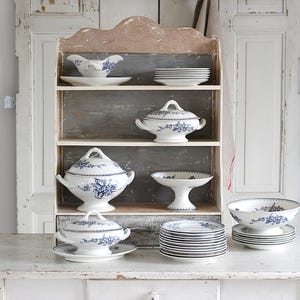 May include: A collection of white porcelain tableware with blue floral designs, including serving bowls, plates, and lidded dishes, displayed on a distressed wooden shelf. The shelf is set against a white background.