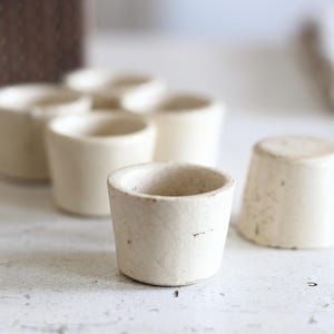 Op de afbeelding: Een verzameling kleine, gebroken witte keramische bekers met een subtiele craquelé glazuurafwerking. Elke beker heeft een eenvoudige, cilindrische vorm met een licht taps toelopende voet en een gladde, open bovenrand, gegroepeerd op een licht oppervlak.
