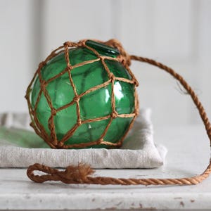 Op de afbeelding: Een groene glazen boei, omhuld door een bruin geweven net, rust op een gevouwen linnen doek. Een bijpassend touw loopt vanaf de bovenkant van de boei en vormt een lus. Het is een decoratief nautisch item.