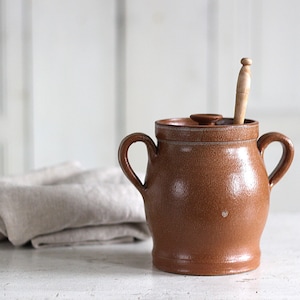 May include: A brown glazed ceramic crock with two side handles and a matching lid. A light-coloured wooden utensil is inserted through a small opening in the lid. The pot has a textured, speckled surface and a rounded body with a flared base.