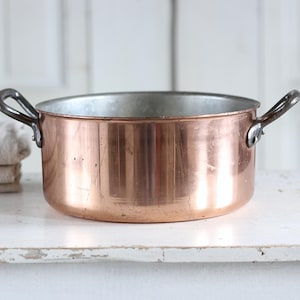 Op de afbeelding: Een koperen kookpot met twee metalen handvatten. De pot heeft een glanzend, reflecterend oppervlak en een zilveren binnenkant. Hij staat op een wit oppervlak, met een stapel gevouwen beige linnen aan de linkerkant.