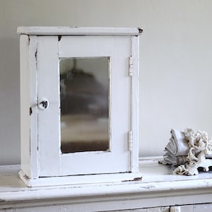 May include: A white wooden wall cabinet with a rectangular mirror panel on the door. The cabinet features a distressed paint finish, a small round knob, and visible metal hinges on the right side. The structure is rectangular with a flat top.