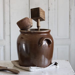 Op de afbeelding: Een donkerbruine geglazuurde keramische pot met twee handvatten aan de zijkant, gevuld met een houten vleeshamer met een getextureerde kop en een houten stamper. Een losse houten lepel ligt ernaast. De pot heeft een gladde, glanzende afwerking.