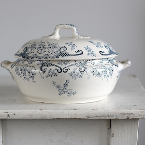 May include: A white ceramic tureen with a lid, adorned with a blue floral and scrollwork pattern. The tureen has two side handles and a handle on the lid. It is placed on a distressed white wooden surface.