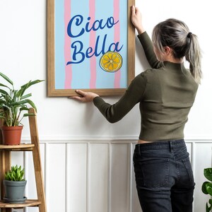 May include: A person hangs a framed print on a white wall. The print features a blue and pink striped background with the text "Ciao Bella" in blue cursive lettering and a yellow lemon slice graphic.