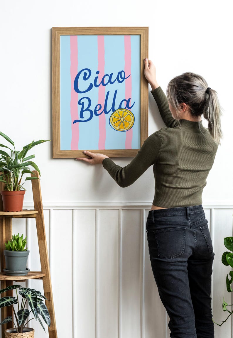 May include: A person hangs a framed print on a white wall. The print features a blue and pink striped background with the text "Ciao Bella" in blue cursive lettering and a yellow lemon slice graphic.