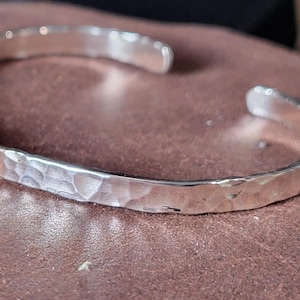 May include: A silver-colored metal cuff bracelet with an open design and a textured, hammered finish. The surface of the bracelet features numerous small, irregular indentations that catch the light, creating a reflective and uneven appearance.