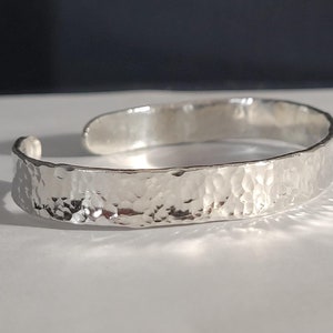 May include: A silver cuff bracelet with a hammered texture. The open-ended design allows for easy wear. The bracelet is approximately 0.5 inches wide.