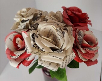 May include: A bouquet of paper roses made from book pages. The roses are a mix of cream and red, with some of the pages featuring black text. The roses are arranged in a vase with green leaves.