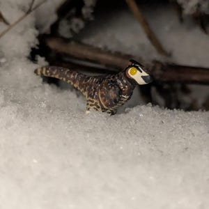 Może przedstawiać: Mała, szczegółowa figurka dinozaura z brązowo-białym nakrapianym ciałem, czarnym grzebieniem i żółtymi oczami, stoi na śniegu. Miniaturowa figurka jest osadzona na tle świeżego śniegu i kilku gałązek.