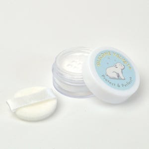 Peut inclure: Un petit pot transparent contenant une poudre blanche, avec un couvercle blanc orné d'un ours polaire de dessin animé et des mots "SILICONE VELVET". Une houppette blanche avec un ruban blanc est à côté du pot. Le produit est sur fond blanc.