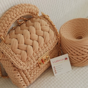 Puede incluir: Un bolso de mano de crochet beige con una correa de cadena dorada y un cierre de llave dorado. Una bola de hilo beige está al lado del bolso.