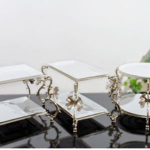 Könnte beinhalten: Drei weiße, mehrstufige Serviertabletts aus Keramik mit silbernen Metallständern. Die Ständer haben ein dekoratives Scroll-Design mit einem Blattmotiv.