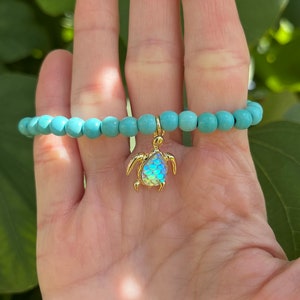 May include: A turquoise beaded bracelet with a gold turtle charm. The turtle's shell is iridescent and has a mermaid-like scale pattern.