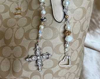 May include: A beige tote bag with a repeating logo pattern. Two beaded charms hang from the handle, one with a rhinestone cross and the other with a rhinestone heart. The beads are white, clear, and gold.