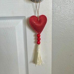 Peut inclure: Ornement en forme de cœur rouge pailleté, orné de trois perles rouges et d'un pompon crème, suspendu à une poignée de porte. L'ornement est suspendu par une ficelle en corde, créant un accent décoratif.