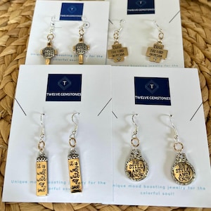 May include: Four pairs of silver and gold earrings with religious and inspirational messages. The earrings feature a cross, a rectangle, and a circle with the words "But first pray", "Bless the Lord oh my soul", "It is well with my soul", and "Be still and know" on them.