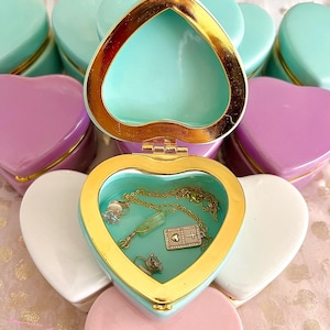 May include: A set of twelve heart-shaped trinket boxes with gold trim. The boxes are in various colors, including pink, white, and light blue. One box is open, revealing a gold necklace and earrings inside.