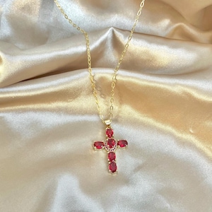 May include: A gold necklace featuring a cross pendant set with red gemstones. The cross design incorporates oval-cut red stones and a central cluster of smaller red gems. The chain is fine, and the pendant is displayed against a satin backdrop.