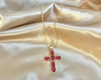 Op de afbeelding: Een gouden ketting met een kruisvormige hanger bezet met rode edelstenen. Het kruisontwerp bevat ovale rode stenen en een centrale cluster van kleinere rode edelstenen. De ketting is fijn, tegen een satijnen achtergrond.