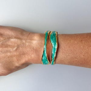 Può includere: Due bracciali rigidi color oro caratterizzati da una forma ondulata e organica. Ogni bracciale presenta un bordo in metallo dorato liscio e lucido e un inserto centrale in resina turchese marmorizzata con finitura lucida.