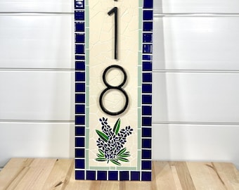 Può includere: Un cartello numerico per la casa in mosaico verticale con il numero 118 in lettere metalliche nere su sfondo bianco. Il mosaico è fatto di piccole piastrelle blu e bianche con un bordo blu. Il cartello ha un disegno di fiore di lupino blu nella parte inferiore.