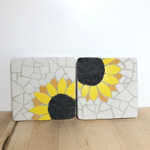 May include: Two white ceramic coasters with a mosaic design featuring black and yellow sunflowers.