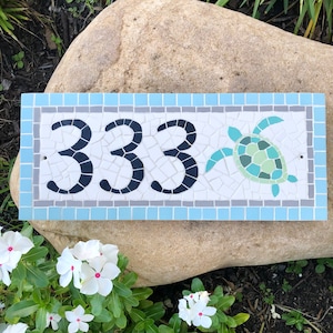 May include: A white mosaic house number sign with the number 333 in black and a blue and green mosaic sea turtle. The sign has a light blue border with a gray inner border.