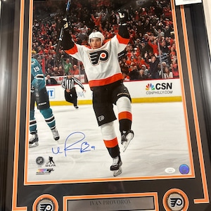 Puede incluir: Una fotografía enmarcada de un jugador de hockey con un jersey blanco y naranja con un logotipo negro y naranja. El jugador levanta su palo en el aire y está rodeado de fanáticos. El nombre del jugador, "Ivan Provorov", está impreso debajo de la imagen. La imagen está firmada por el jugador.