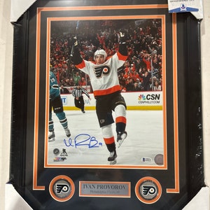 Puede incluir: Una foto enmarcada de un jugador de hockey con una camiseta blanca y naranja con el número 9 en la parte delantera. El jugador levanta los brazos en señal de celebración. El nombre del jugador, Ivan Provorov, y el nombre del equipo, Philadelphia Flyers, están impresos debajo de la foto. La foto está firmada por el jugador.