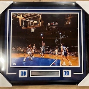 Peut inclure: Photographie encadrée d'un match de basket-ball. L'image montre des joueurs en action, avec un joueur tirant un ballon. Le cadre a un passe-partout bleu et un cadre noir, avec la signature du joueur. Le cadre est rectangulaire.